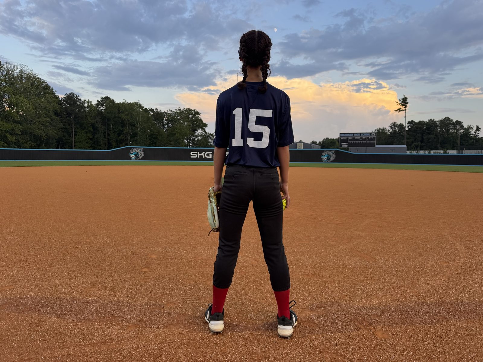 An ORIGIN athlete in a navy shirt numbered 15 stands on the infield at Seckinger High School at dusk, glove in one hand and a ball in the other.